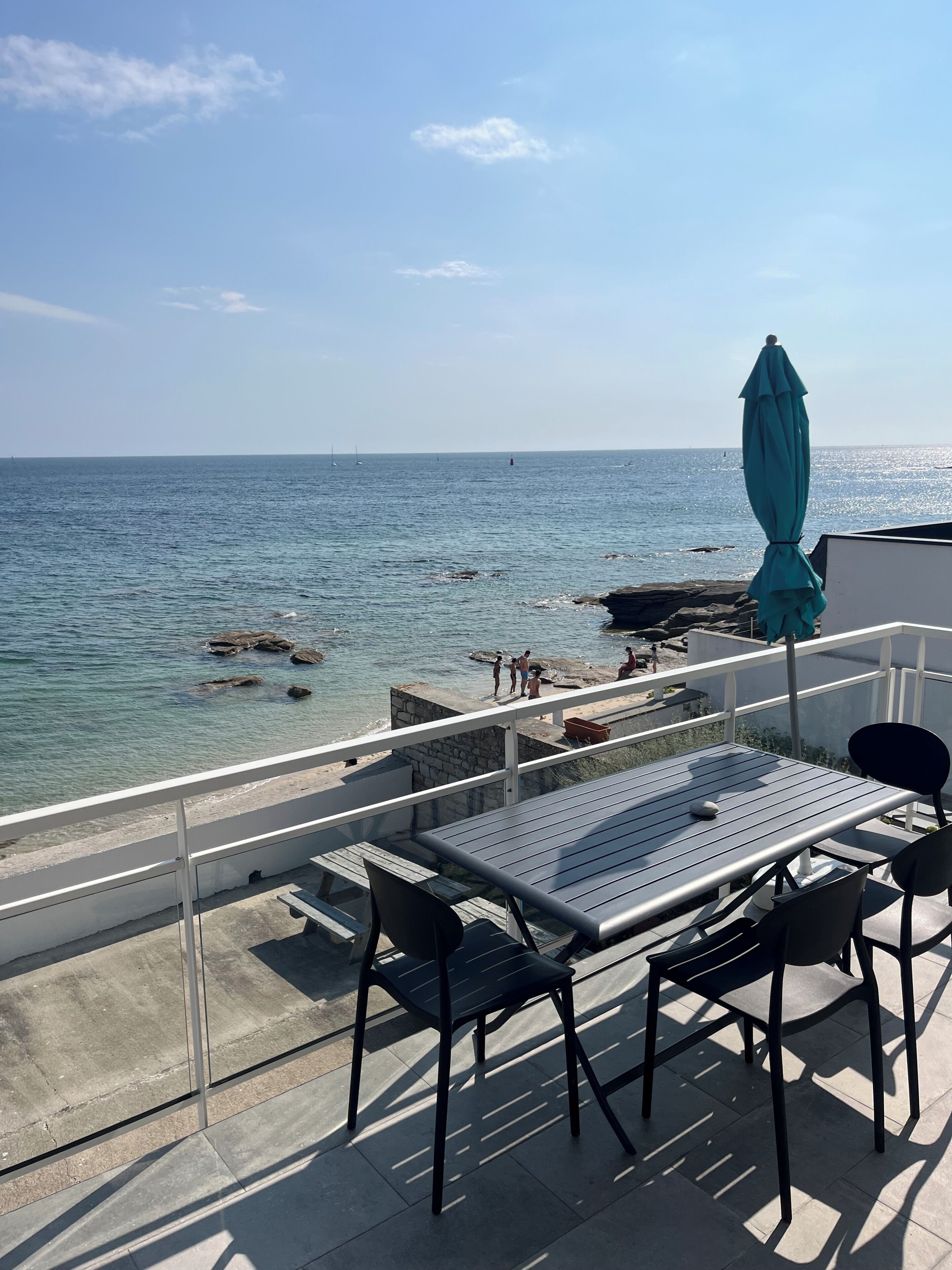 A balcony with a table and chairs overlooking a rocky coastline with the sea.