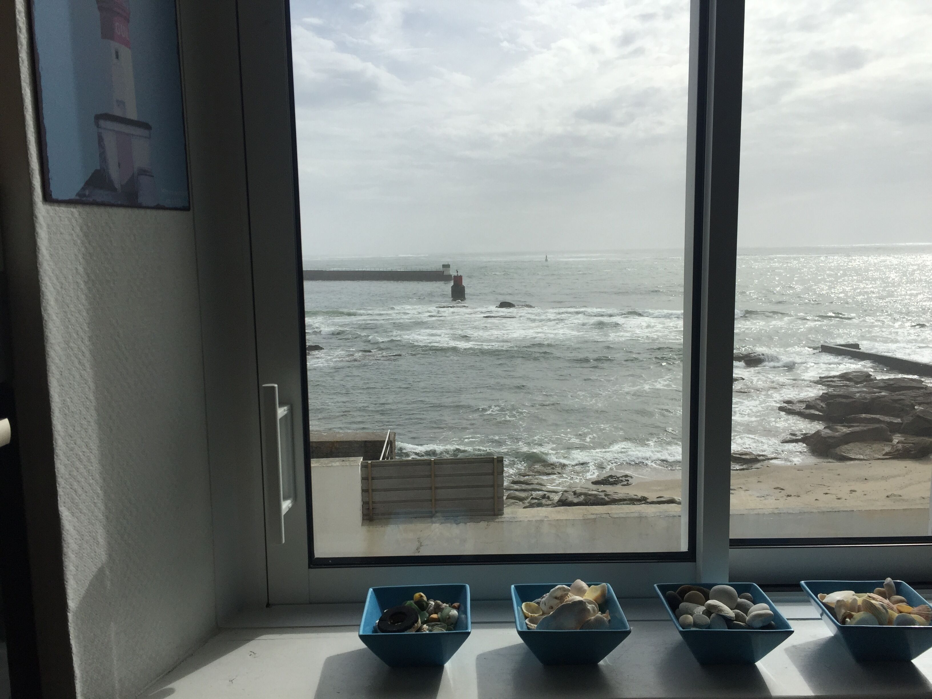 A view of the sea through a window with four blue bowls of seashells on the windowsill.