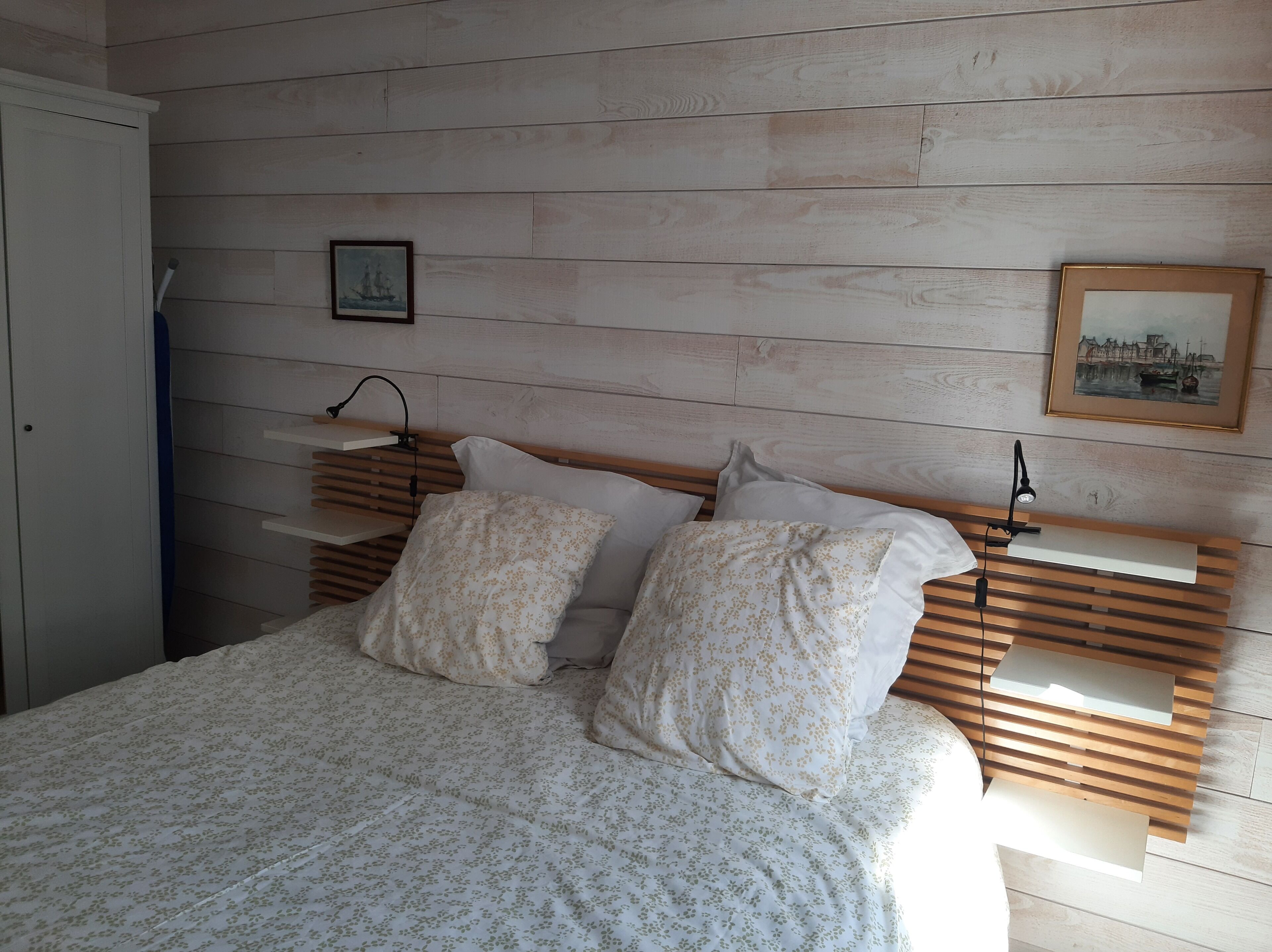 A bedroom with a wooden headboard, two pillows, a bedside table, and a wall-mounted lamp.