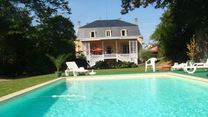 En swimmingpool med kristallklart blått vatten, omgiven av vita solstolar och ett hus i bakgrunden.