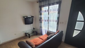 A living room with a dark brown sofa, a flat-screen TV mounted on the wall, and a window with curtains.