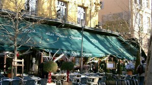 Un café al aire libre con toldo verde, mesas en la terraza y un edificio de arquitectura clásica al fondo.