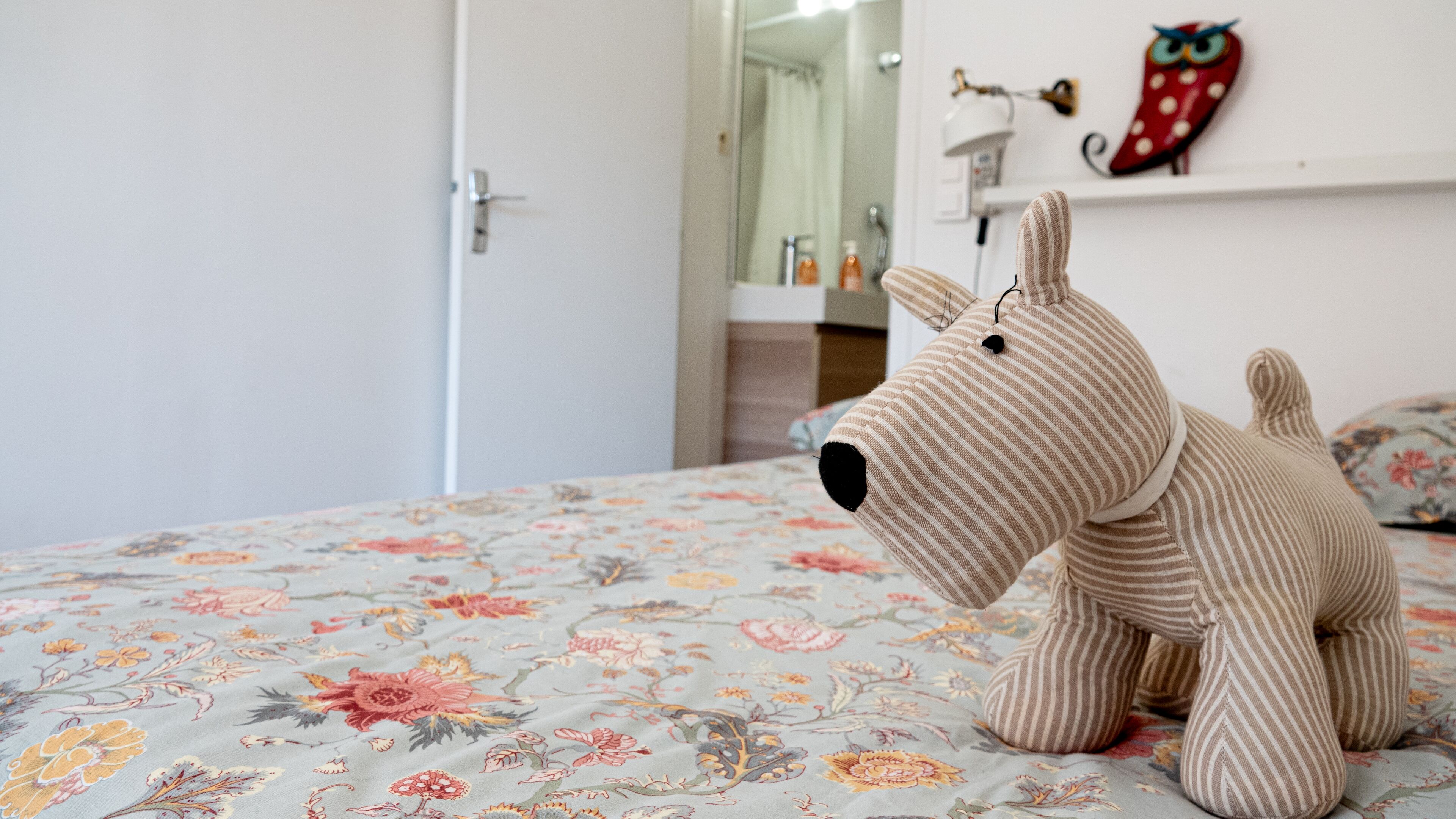 Ein Bett mit einem blumigen Überwurf, ein gestreiftes Stofftier auf dem Bett, eine weiße Tür und ein Spiegel mit einem roten und weißen gepunkteten Gegenstand an der Wand.