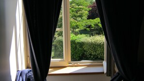 Ein Fenster mit dunklen, halb geöffneten Vorhängen, durch das sich Grünblick zeigt.
