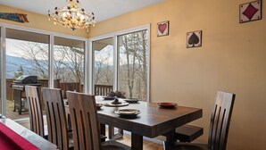 Un espace repas comprenant une table et des chaises en bois, un lustre et de grandes fenêtres offrant une vue sur les arbres et les montagnes.
