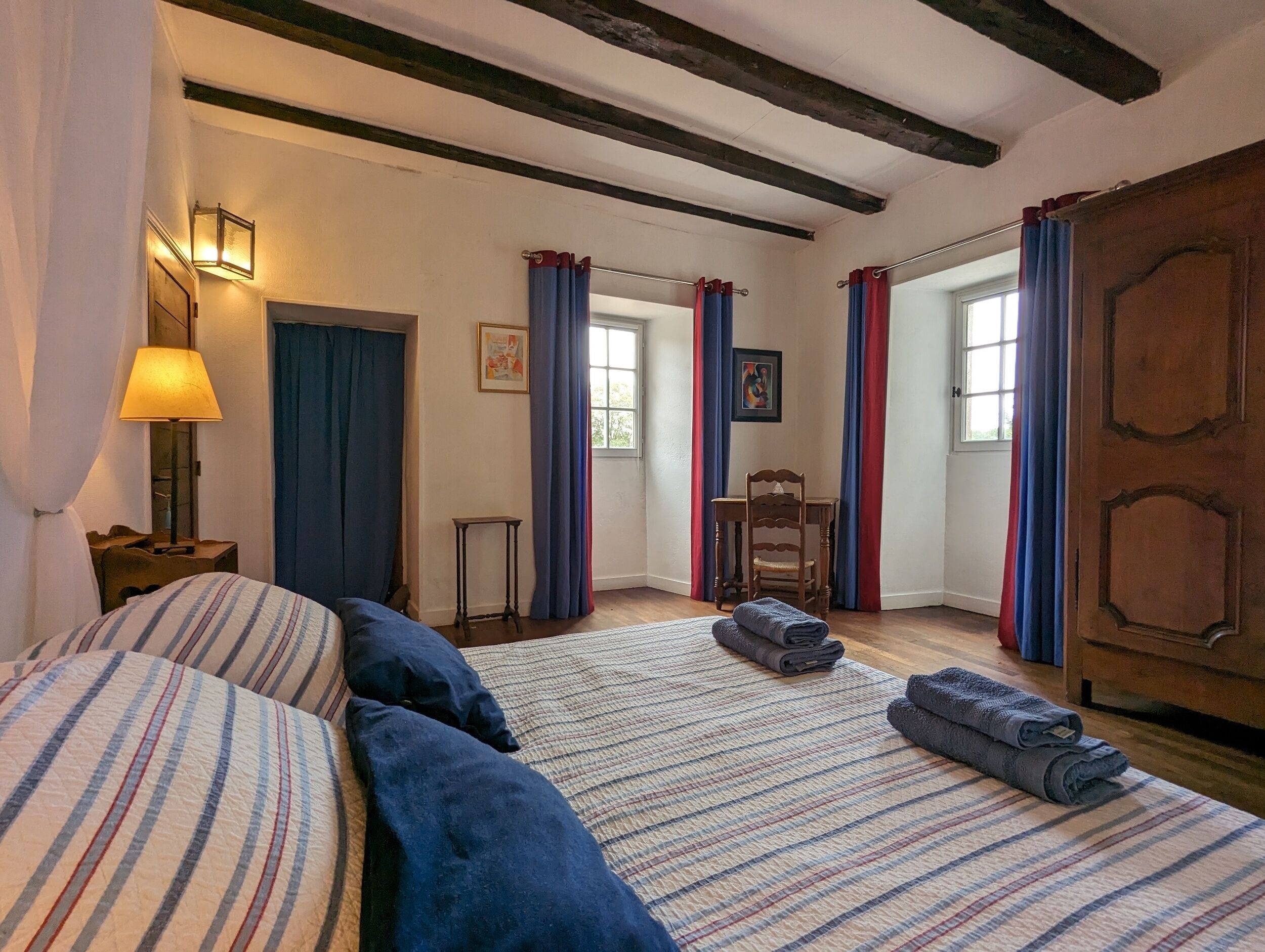 Een knus vertrek met een bed, een tafel en een stoel. Gordijnen, een lamp en een deur zijn aanwezig.