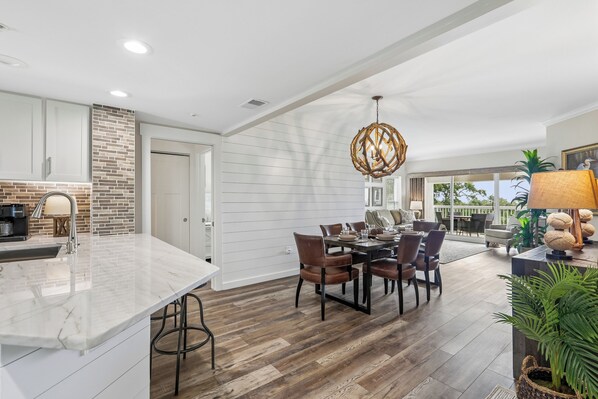 A spacious kitchen with a marble countertop, a dining area with a wooden table and chairs, and a large window offering a view of the outdoors.