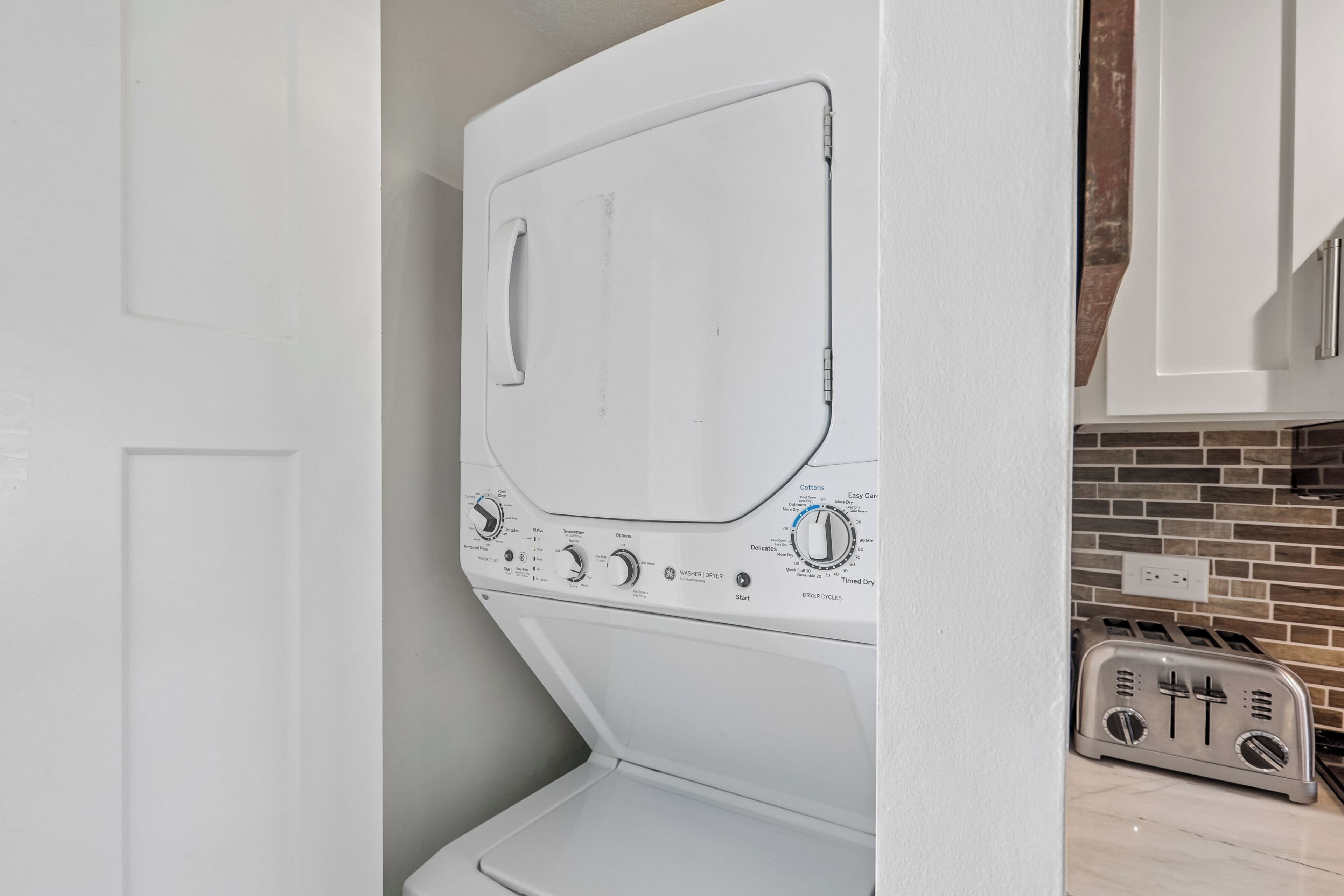 A front view of a white front-loading washing machine with control panel, located in a kitchen area with a brick backsplash and a toaster.