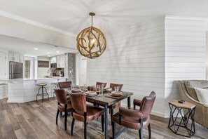 Une cuisine moderne dotée d’un îlot central, un coin repas avec une table en bois et des chaises en cuir, et un salon avec un canapé et une table d’appoint.