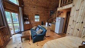 Interior de una cabaña con suelo de madera, un sofá azul con cojines, una zona de cocina con refrigerador y una mesa de comedor.
