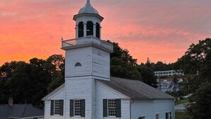 En vit kyrka med ett klocktorn och en vindflöjel, omgiven av träd och ett bostadsområde.