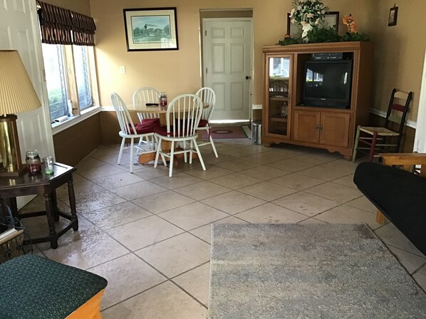 Ein Wohnzimmer mit Esstisch, weißen Stühlen, einer Couch, einem Fernseher und einem Schrank.