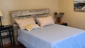 Een slaapkamer met een houten hoofdbord, een bed met een lichtblauwe spreideken, twee kussens, een nachtkastje met een lamp en een potplant.