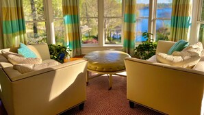 A cozy room with two cream-colored armchairs, a round ottoman, and a view of a lake through large windows.