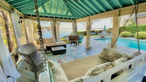 A covered outdoor seating area with a swing chair, a sofa, and a pool in the background.