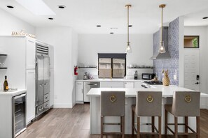 Cocina moderna con armarios blancos, una isla central con taburetes de bar, electrodomésticos de acero inoxidable e iluminación colgante.