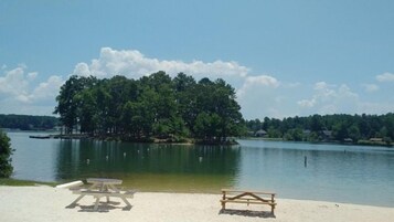 Vista sobre un lago con playa arenosa, dos mesas de picnic y un muelle que se adentra en el agua.