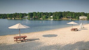 Una playa arenosa con mesas de picnic y sombrillas, un lago tranquilo y casas al fondo.
