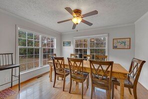 Une salle à manger avec une table et des chaises en bois, de grandes fenêtres et un ventilateur de plafond.