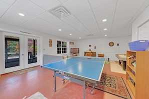 Une pièce comprenant une table de ping-pong, un bureau, une chaise, une bibliothèque et une porte donnant sur un balcon.