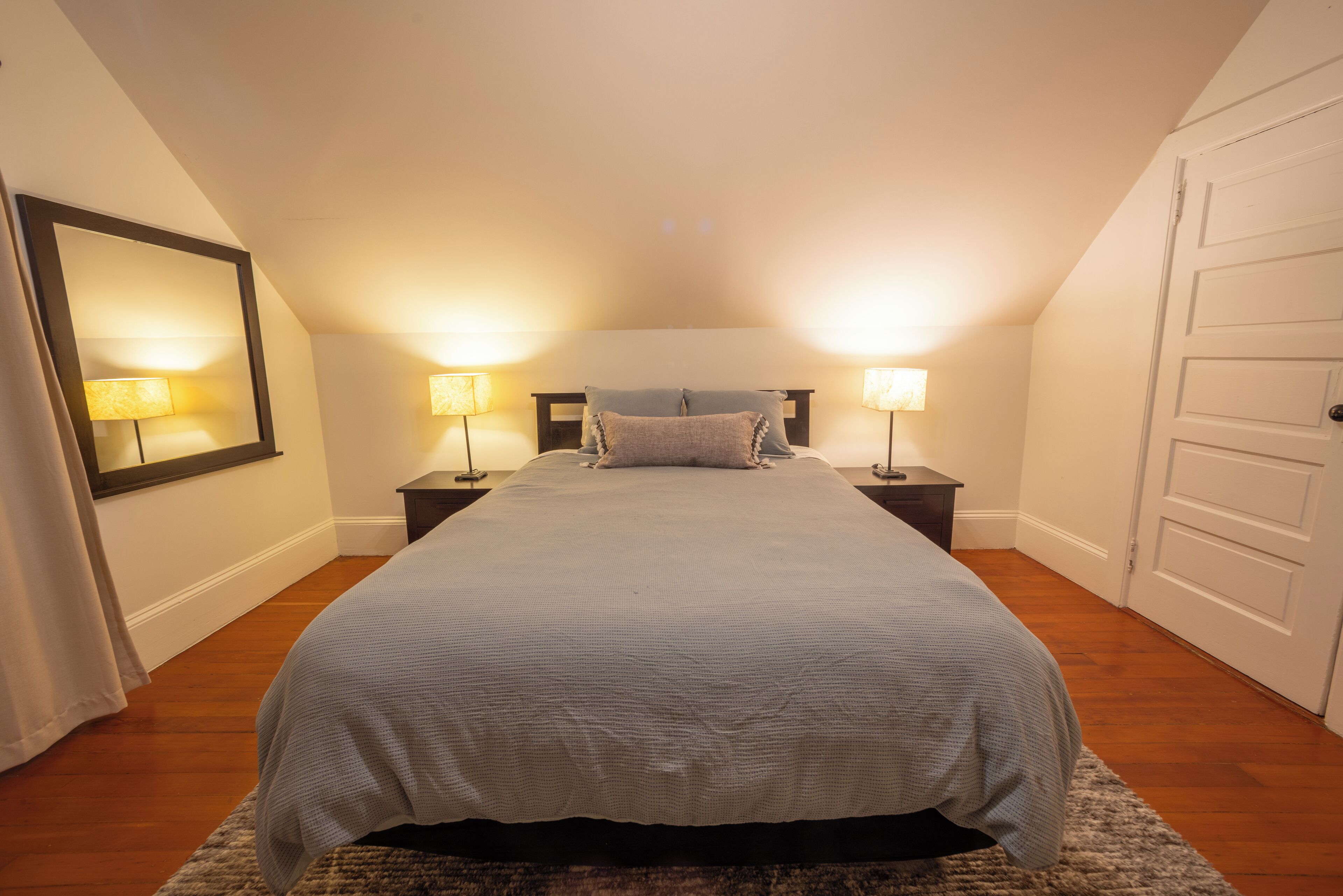 A bedroom with a large bed, two bedside lamps, a mirror, and a door.