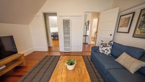 Una sala de estar con un televisor sobre un mueble de madera, un sofá azul con un cojín estampado, una mesa de centro de madera con una planta y una alfombra de cuadros.