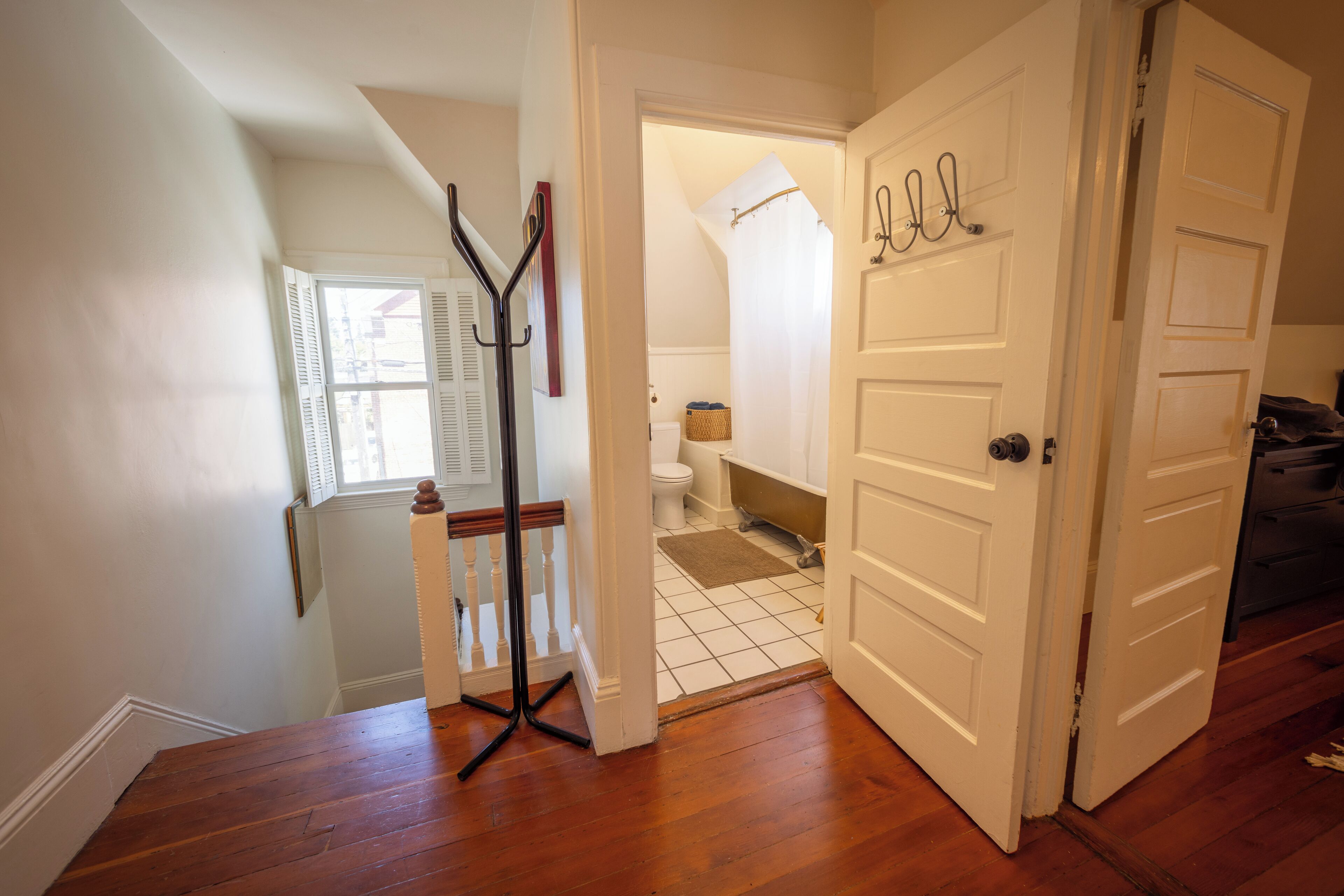 A hallway with wooden flooring, a white door, and a view into a bathroom with a toilet, bathtub, and a shower curtain.