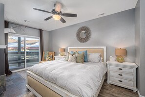A bedroom with a bed, two nightstands, a ceiling fan, a window with a view, and a decorative wall piece.