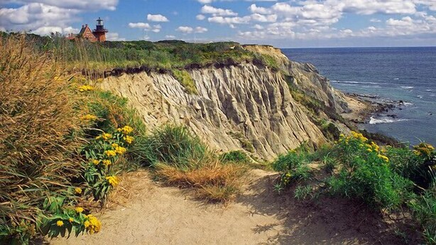 A coastal landscape with a cliff, yellow flowers, and a lighthouse in the background. - Stunning Sunsets on Quonnie Waterfront- 2 Kayaks, Beach Pass & Backyard Clamming (Charlestown)