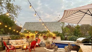 Un patio trasero con una parrilla, piscina, mobiliario exterior e iluminación con luces colgantes.