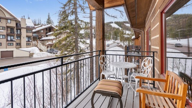 A wooden balcony with a table and chairs, overlooking a snowy landscape with buildings and trees.