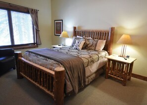 A bedroom with a wooden bed frame, two bedside tables with lamps, a chair, and a window with blinds. - The Pines 3 Bedroom Condo (Whitefish)