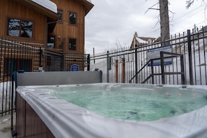 A wooden building with a hot tub in front, surrounded by a metal fence. - The Pines 3 Bedroom Condo (Whitefish)