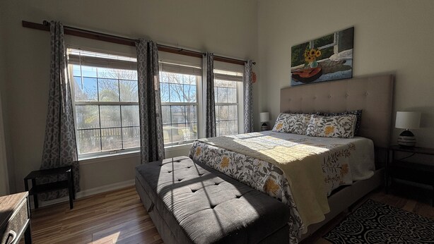 A bedroom with a bed, a bench, two windows with curtains, a painting, and a bedside table with a lamp. - 1 Bedroom Studio located near Isle of Palms and downtown Charleston! (Mt Pleasant)