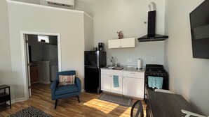 A compact kitchen with a refrigerator, stove, and cabinets. A blue chair and a small table are in the foreground. - 1 Bedroom Studio located near Isle of Palms and downtown Charleston! (Mt Pleasant)