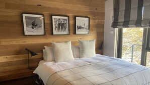 Une chambre à coucher avec une tête de lit en bois, un lit recouvert d’une couette à carreaux, trois tableaux encadrés accrochés au mur et une fenêtre donnant sur la forêt.