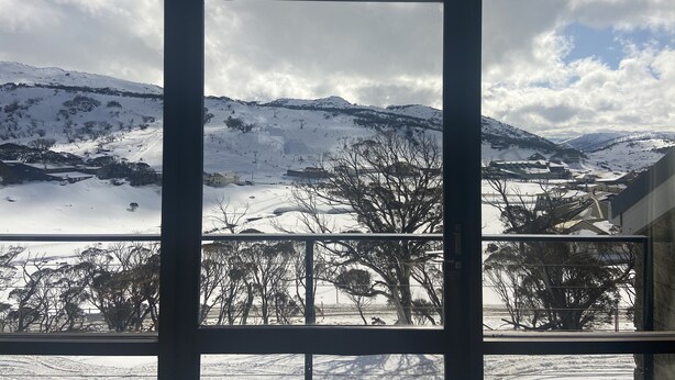 Une vue à travers une fenêtre sur un paysage enneigé, avec des montagnes, des arbres et un bâtiment au premier plan.