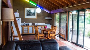 A cozy living area with a sofa, wooden dining table, and chairs, a kitchen with a refrigerator, and large sliding glass doors to a garden. - Back Forest Retreat Sunrise Cottage (Back Forest)