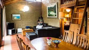 A cozy living room with a wooden ceiling, a black leather sofa, a dining table with chairs, a TV, and a fireplace. - Back Forest Retreat Sunrise Cottage (Back Forest)