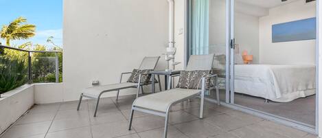 A balcony with two chairs and a table, a bed with a white bedspread, and a view of palm trees.
