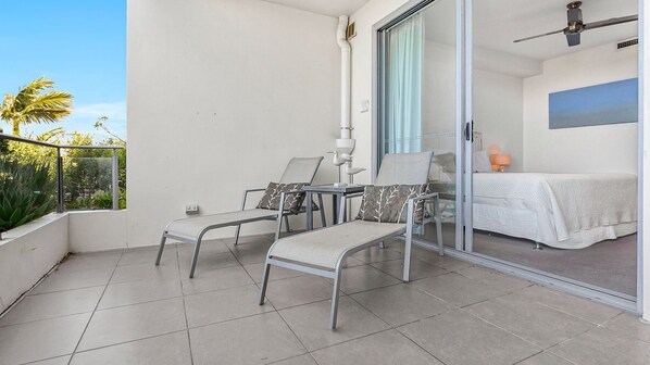 Un balcon avec deux chaises et une table, un lit recouvert d’une courtepointe blanche, et une vue sur des palmiers.