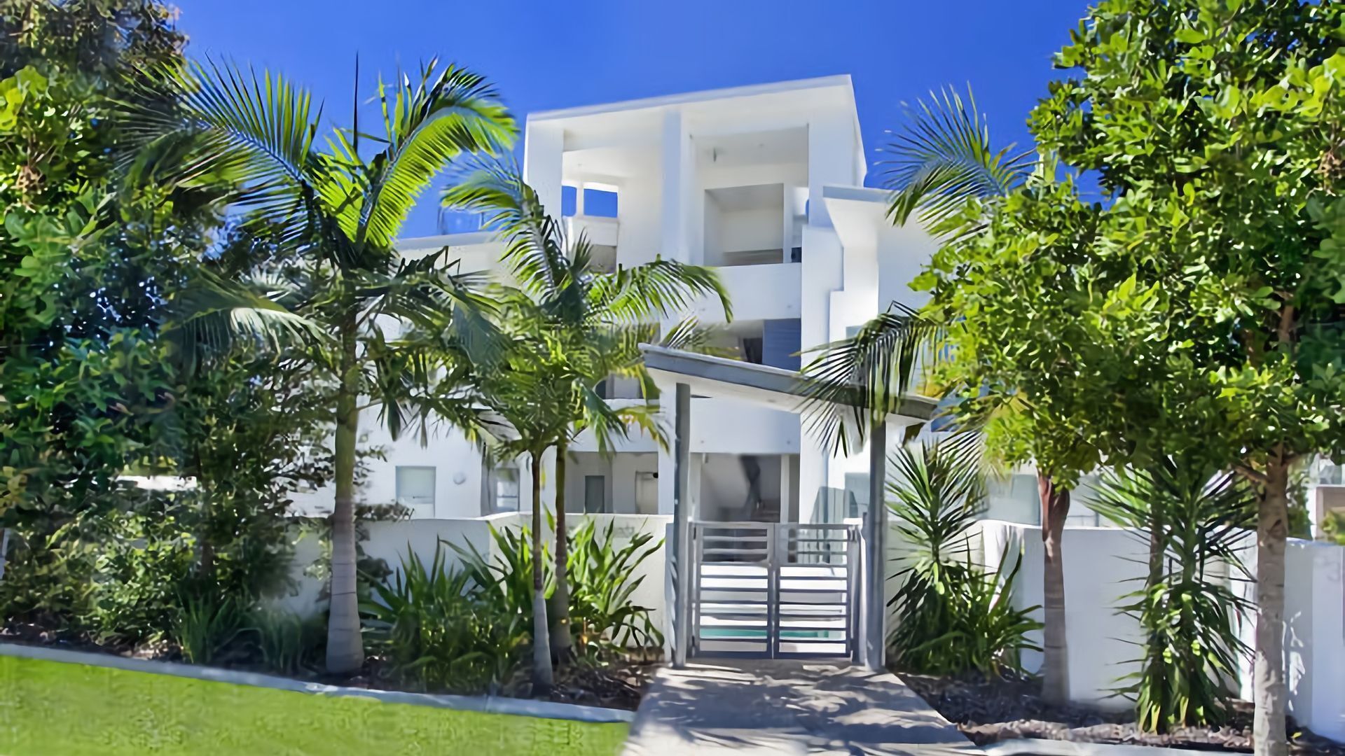 A modern multi-story building with a white facade, surrounded by lush greenery and palm trees.