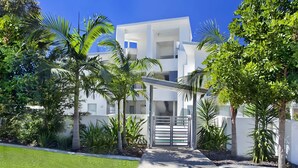 A modern multi-story building with a white facade, surrounded by lush greenery and palm trees.