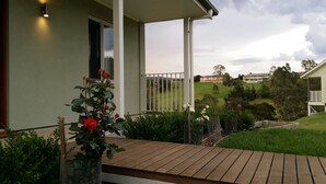 A wooden deck with a railing, a potted rose bush, and a view of a grassy field and buildings in the distance. - Highfields Country group Cottage  2 Bedroom (Kurrajong)