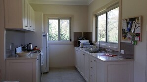 A compact kitchen with white cabinets, a microwave, a kettle, and a sink. There are two windows providing natural light, and a bulletin board with various notices. - Highfields Country group Cottage  2 Bedroom (Kurrajong)