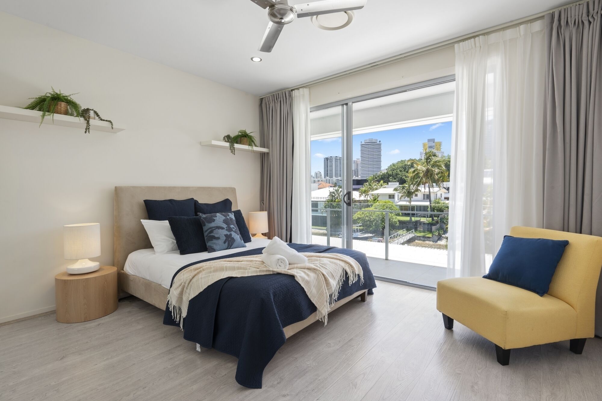 A modern bedroom with a bed, bedside tables, a chair, and a balcony with a city view.