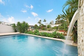 Piscina com cascata, rodeada por plantas tropicais e palmeiras, com edifícios ao fundo.