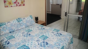 Une chambre à coucher avec une couvre-lit à motifs, une table de chevet avec une lampe et une porte donnant sur un coin cuisine.