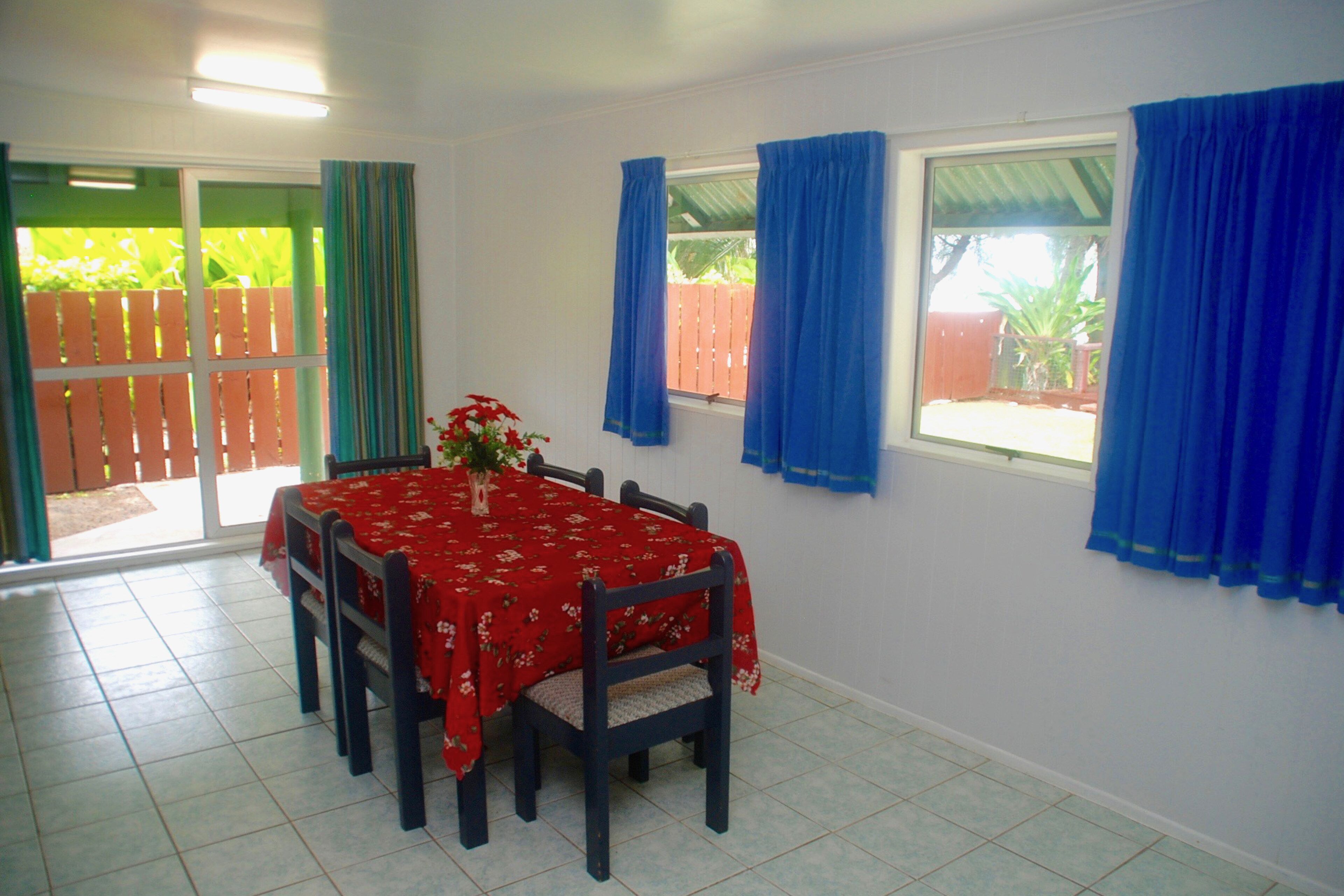 Uma área de refeições com uma mesa posta para seis pessoas, um vaso de planta e vista para uma cerca de madeira e vegetação lá fora.