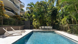 A swimming pool with lounge chairs and a table, surrounded by lush greenery and a multi-story building.